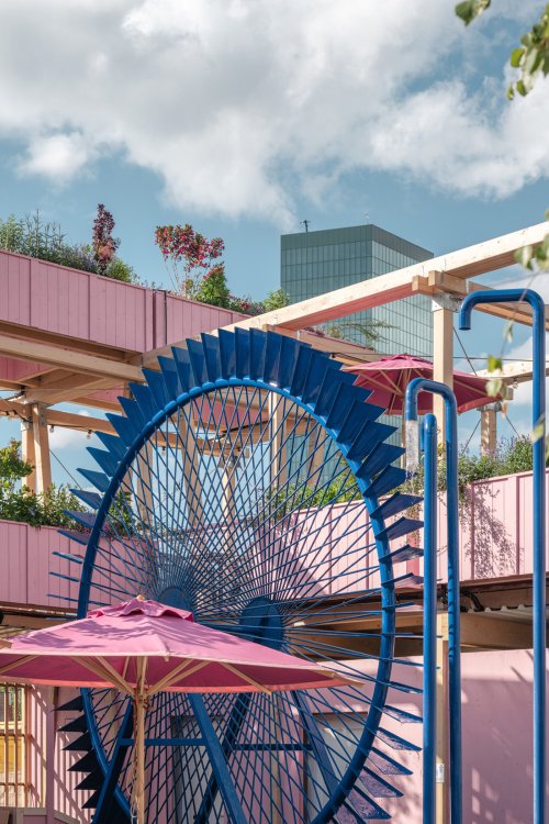 Ein grosses, blaues Metall-Wasserrad als spielerisches Element in der Remise Rosa in Zürich West. Es ist vor einer rosa Holzwand und unter hellen Holzbalken platziert, daneben sind blaue Wasserrohre und rosa Sonnenschirme zu sehen.