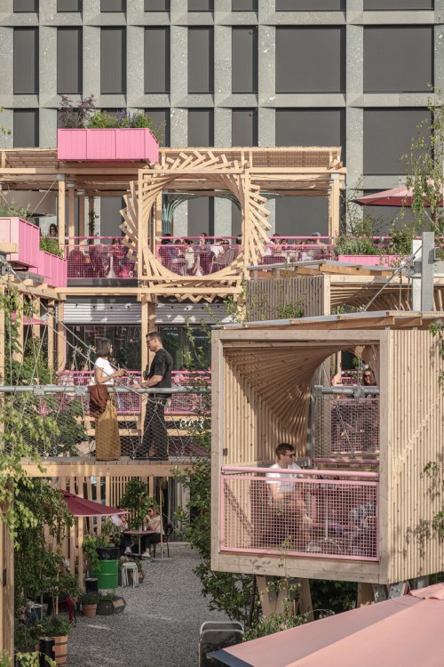 Detaillierte Ansicht der mehrgeschossigen Holzstruktur der Remise Rosa: Menschen unterhalten sich auf einer Seilbrücke und in kleinen, erhöht liegenden Holzkabinen. Im Hintergrund sind weitere Terrassenebenen mit rosa Akzenten und die graue Fassade eines Bürogebäudes zu sehen.