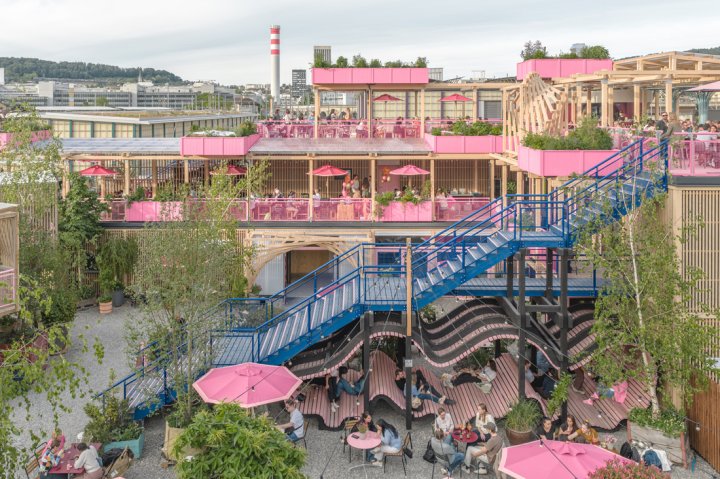 Gesamtansicht der Remise Rosa in Zürich West bei Tag: Eine belebte, mehrgeschossige Holzkonstruktion mit rosa Terrassen, blauen Treppen und wellenförmigen Sitzbänken im Erdgeschoss. Zahlreiche Gäste nutzen die verschiedenen Ebenen und Sitzbereiche des urbanen Treffpunkts.