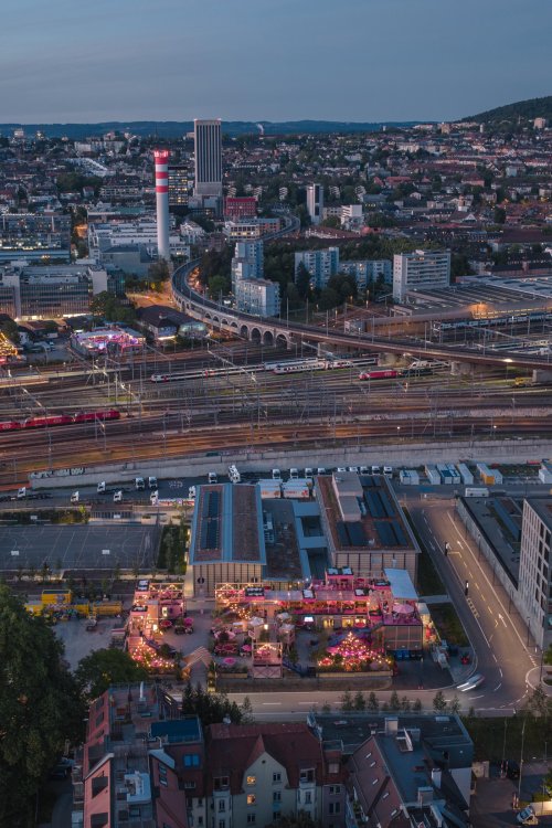 Nächtliche Luftaufnahme von Zürich West im Hochformat: Die hell erleuchtete, rosafarbene Remise Rosa bildet einen leuchtenden Kontrast zu den angrenzenden dunklen Bahngleisen und dem urbanen Panorama der Stadt bei Nacht.