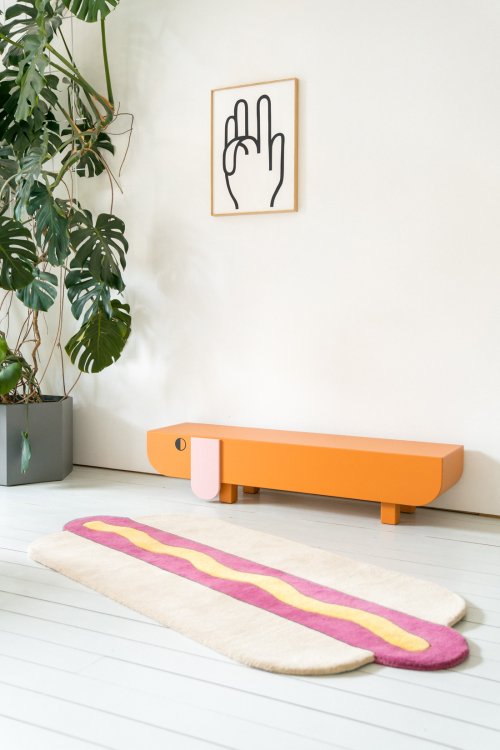 Minimalistisches Interieur mit einer orangefarbenen Sitzbank in Form eines Dackels vor einer weissen Wand.