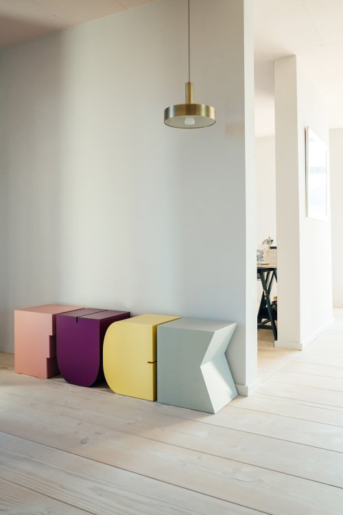Vier bunte Design-Hocker in Form der Buchstaben F, U, C und K sind nebeneinander an einer weissen Wand aufgereiht und bilden das Wort „FUCK“. Die Buchstaben-Möbel in den Farben Rosa, Lila, Gelb und Hellgrau stehen in einem minimalistisch eingerichteten Raum mit hellem Holzboden unter einer modernen, goldfarbenen Pendelleuchte.