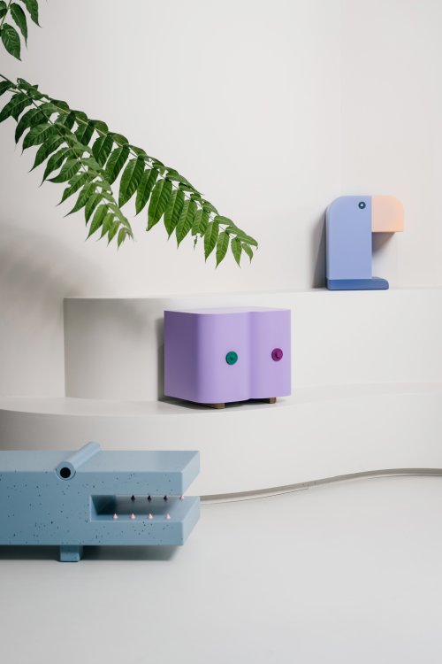 Minimalistische Interieur-Szene mit verspielten Designmöbeln: ein blauer Krokodil-Hocker im Vordergrund, ein lilafarbener Beistelltisch in Form stilisierter Brüste auf einer Stufe sowie ein kleiner Tukan-Hocker an der Wand. Eine grosse grüne Pflanze ragt in den hellen, weissen Raum hinein.