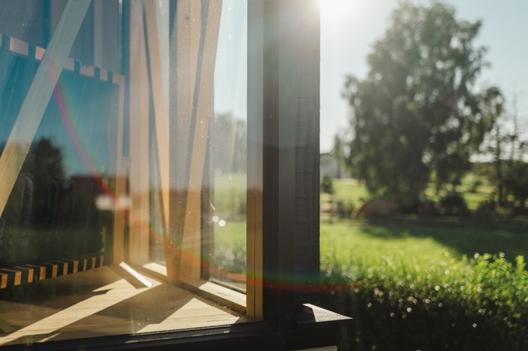 Nahaufnahme der Glasecke des Pavillons im Gegenlicht. Hinter der Glasscheibe ist die hölzerne Fachwerkkonstruktion mit diagonalen Streben erkennbar. Im Hintergrund erstreckt sich eine sonnige, grüne Landschaft.