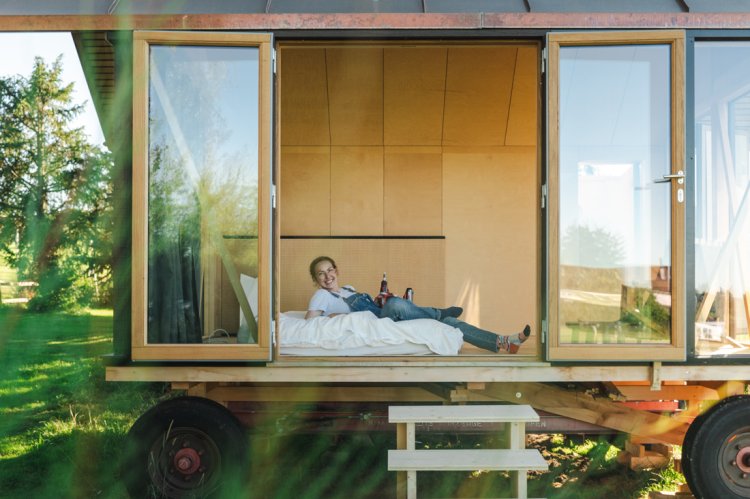 Architektin Eliane Windlin liegt lächelnd auf einer Matratze in einem kleinen Holzpavillon auf Rädern. Die grossen Flügeltüren an der Front stehen offen und geben den Blick auf den hellen Innenraum frei. Der Pavillon steht auf einer Wiese und ist von Bäumen umgeben.
