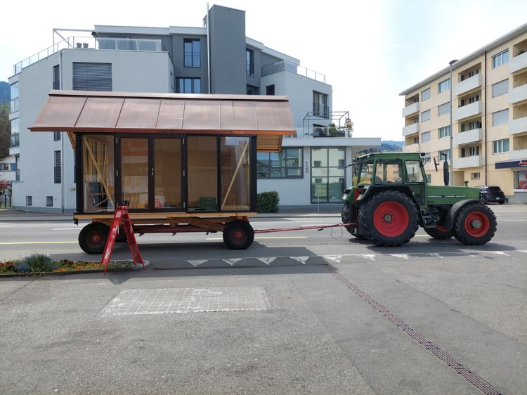 Ein Traktor zieht den mobilen Holzpavillon auf einem Fahrgestell durch eine Ortschaft. Die grossen Glasflächen und das kupferverkleidete Giebeldach sind gut sichtbar, im Hintergrund stehen moderne Wohngebäude.