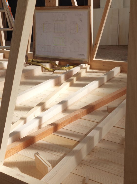 Nahaufnahme der hölzernen Bodenkonstruktion des Pavillons im Rohbau. Im Hintergrund ist ein technischer Konstruktionsplan auf einem Klemmbrett zu sehen, der an dem hellen Eschenholz-Fachwerk befestigt ist. Sonnenlicht fällt schräg auf die Holzbalken.