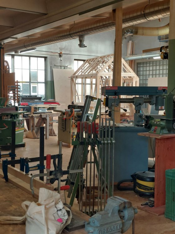 Blick in eine belebte Schreinerwerkstatt mit verschiedenen Holzbearbeitungsmaschinen, Werkzeugen und Schraubzwingen im Vordergrund. Im Hintergrund ist das helle Holzskelett (Rohbau) des Pavillons mit seiner charakteristischen Giebelform zu sehen.