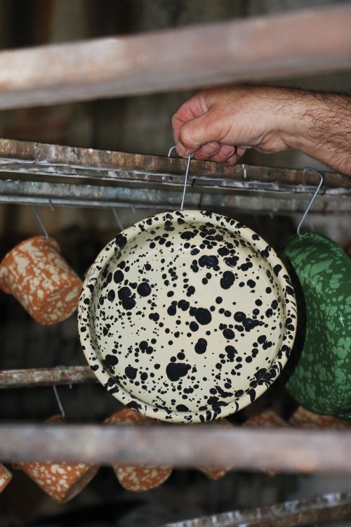Detailaufnahme aus der Manufaktur: Ein behaarter Männerarm hängt einen beige-schwarz gesprenkelten Emaille-Teller an einem Metalldraht in ein Trocknungs- oder Brenngestell. Im Hintergrund sind weitere aufgehängte Becher und Schalen in Terrakotta und Grün erkennbar.