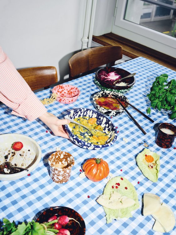 Ein reich gedeckter, hellblau-weiss karierter Tisch mit verschiedenen gesprenkelten Emaille-Schalen und -Bechern von ÉMAÉ STUDIO in Blau, Rot, Braun und Schwarz. In den Schalen befinden sich Reste von Speisen wie Salat, Suppe und Granatapfelkernen. Eine Hand im rosa-weiss gestreiften Hemd greift nach einer blauen Schale.