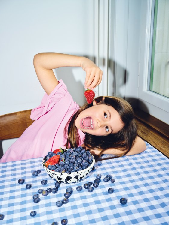 Ein Mädchen in einem rosa T-Shirt lehnt lachend den Kopf auf einen blau-weiss karierten Tisch und hält sich eine Erdbeere vor den weit geöffneten Mund. Vor ihr steht eine schwarz-weiss gesprenkelte Emaille-Schale (Mirari Bowl) von ÉMAÉ STUDIO, die hoch mit Heidelbeeren und Erdbeeren gefüllt ist. Weitere einzelne Heidelbeeren liegen auf dem Tisch verteilt.