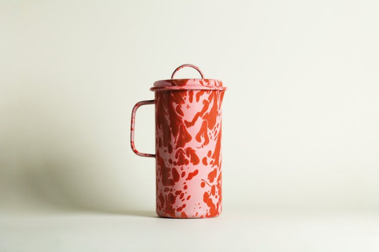 Studio-Produktfoto einer zylindrischen Emaille-Karaffe mit Deckel von ÉMAÉ STUDIO. Das Muster besteht aus lebendigen, fliessenden Farbspritzern in leuchtendem Rot (Tomato) auf einem hellrosa Hintergrund (Peony). Cleane, minimalistische Ästhetik vor einem neutralen, hellen Hintergrund.