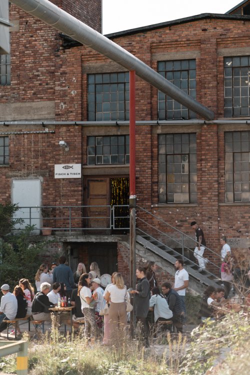 Besucherinnen und Besucher sitzen und stehen bei einem Event vor der Backsteinhalle von Fisch&Tisch in Aßlar. Über dem Eingang ist das Logo des Möbellabels zu sehen; grosse Industrie-Fenster und ein silbernes Lüftungsrohr prägen die Fassade.