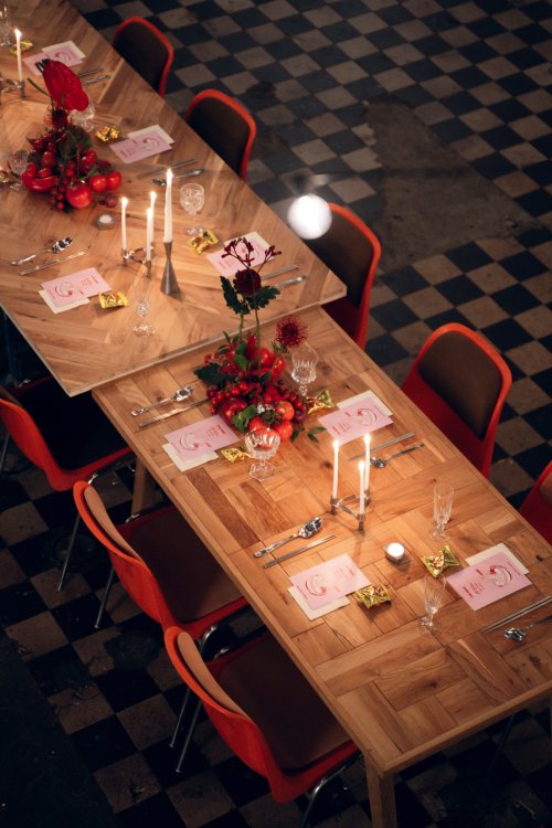 ine lange, festlich gedeckte Tafel aus Fischgrätparkett-Tischen in einer Industriehalle mit schwarz-weiss kariertem Boden. Die Tische sind mit brennenden Kerzen in silbernen Leuchtern, rotem Blumenschmuck, Weingläsern und roten Designerstühlen arrangiert.