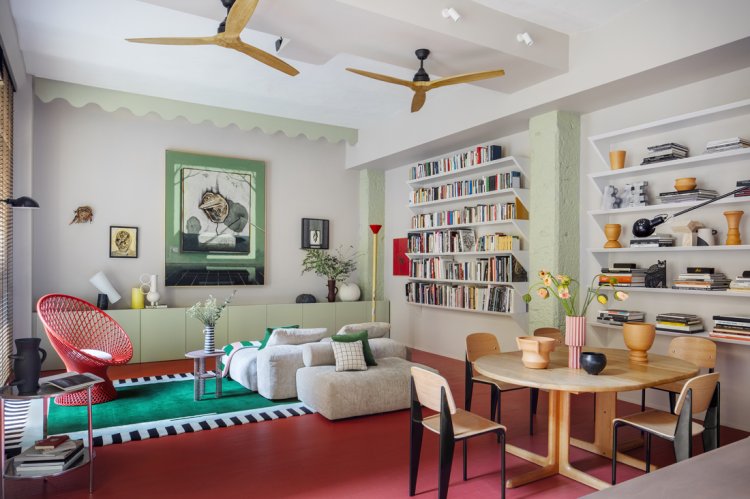Helles, farbenfrohes Wohnzimmer mit Bücherregalen, Esstisch aus Holz, roten und grünen Teppichen sowie zwei Deckenventilatoren.