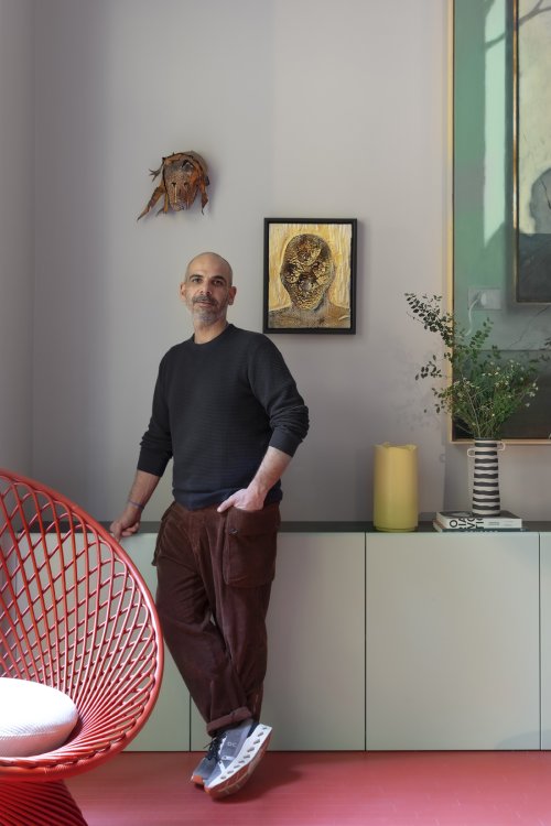 Ein Mann mit Bart in einem dunklen Pullover und braunen Cordhosen lehnt in einem modern gestalteten Wohnraum an einem hellgrünen Sideboard. Hinter ihm hängen abstrakte Kunstwerke an der weissen Wand, während neben ihm eine gestreifte Vase mit Eukalyptuszweigen und ein gelbes Dekoelement stehen. Im Vordergrund setzt ein markanter roter Korbsessel einen farblichen Akzent auf dem ebenfalls roten Boden.