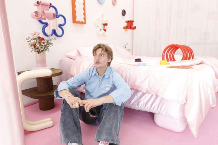 Gustaf Westman sitzt in einem pastellrosa eingerichteten Raum auf dem Boden vor einem Bett und blickt nach oben; um ihn herum stehen bunte, skulpturale Designobjekte.