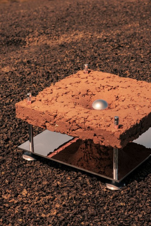 Gravity Well: Stampflehm-Objekt auf einer dunklen, geröllartigen Oberfläche, mit einer runden Vertiefung und einer kleinen Metallkugel in der Mitte, getragen von einer reflektierenden Edelstahlkonstruktion.