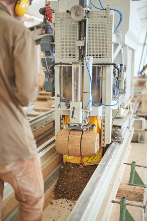 Ein Mitarbeiter in Arbeitskleidung und Gehörschutz beobachtet eine Maschine, die grosse, runde Lehm-Deckensteine presst und auf eine Palette schiebt. Ein Haufen lockerer Lehm liegt auf dem Förderband unter der Maschine.
