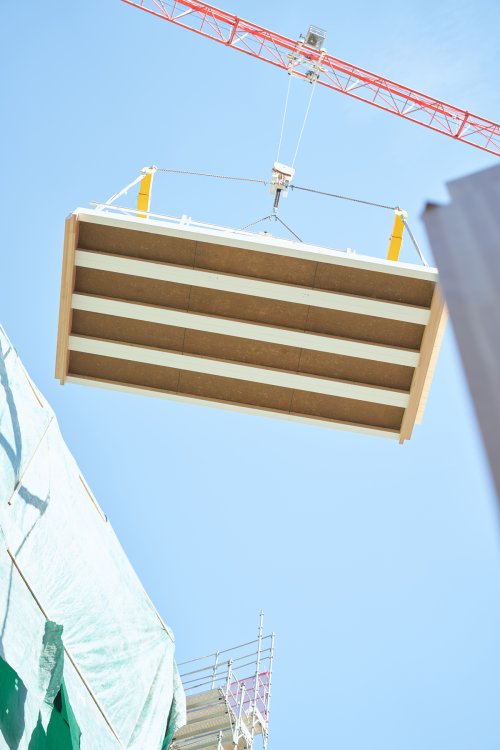 Blick von unten auf einen roten Baukran, der ein grosses, vorgefertigtes Deckenelement aus Holz und Lehm an Seilen hochhebt. Im Vordergrund sind eine grüne Schutzplane und ein Teil eines Baugerüsts zu sehen.