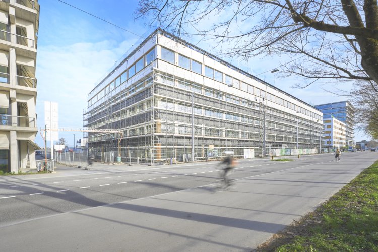 Aussenansicht eines grossen, modernen Bürogebäudes im Bau. Die Holz- und Glasfassade ist fast vollständig hinter einem Metallgerüst verborgen. Das Gebäude erstreckt sich entlang einer breiten Strasse mit Velowegen, auf der eine Radfahrerin vorbeifährt