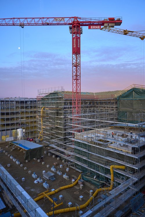Blick von oben auf die Baustelle des HORTUS-Gebäudes in der Dämmerung mit einem grossen roten Baukran im Zentrum. Der Rohbau ist von Gerüsten umgeben und gelbe Belüftungsschläuche ziehen sich über das Gelände vor dem Gebäude.