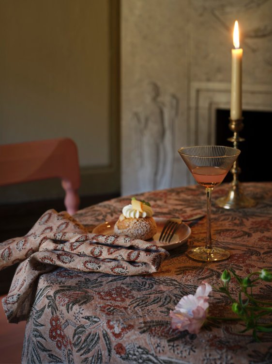 Stilvoll gedeckter Tisch mit floral gemusterter Tischdecke in warmen Rosé-, Braun- und Grüntönen, dazu ein Dessert, ein Cocktailglas und eine Kerze in stimmungsvollem Licht.