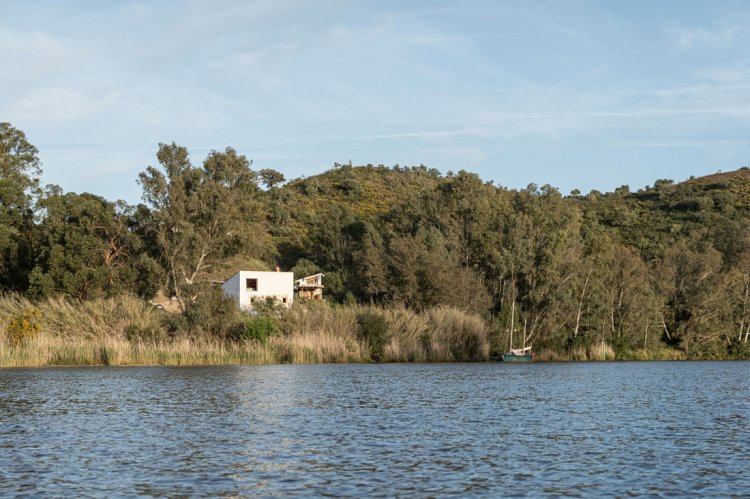 Haus Stone and Water am Ufer des Guadiana mit Boot vor Uferlandschaft mit Bäumen und Schilf.