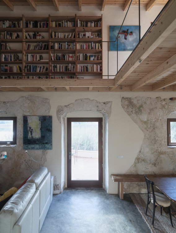 Innenraum von Stone and Water mit offener Galerie aus Holz, grossem Bücherregal, freigelegten Steinmauern und Blick durch die Tür auf die Terrasse.