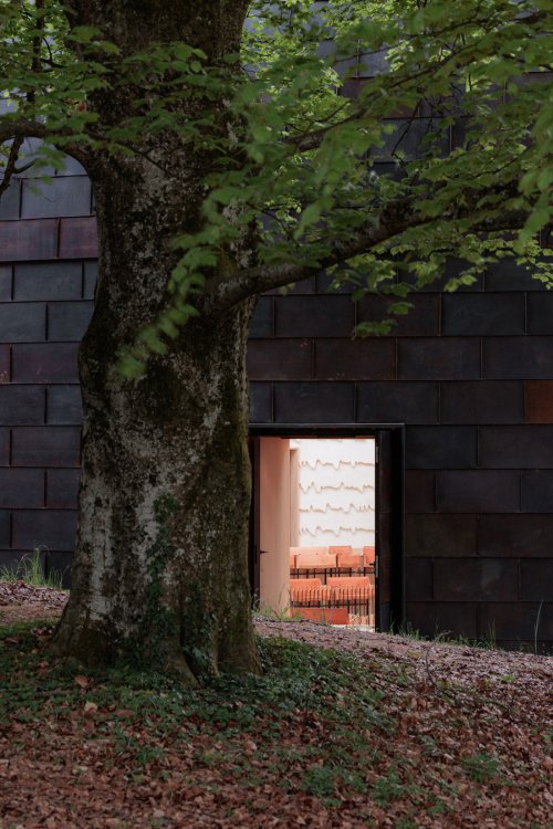 Ein grosser Baum steht vor der dunklen, kupferverkleideten Fassade von «La Source Vive». Durch eine offene Tür sind der helle Saal, die orangefarbenen Sitzplätze und die strukturierte Wandoberfläche zu sehen.