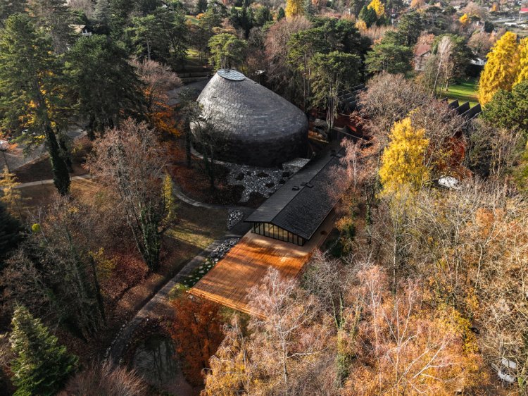 Luftaufnahme von «La Source Vive» mit seiner rundlichen, kupfergedeckten Dachform und dem angrenzenden Foyer mit Holzterrasse, eingebettet in einen herbstlich gefärbten Wald.