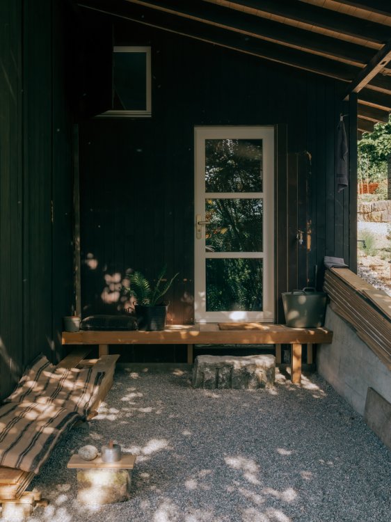 Eingangsbereich des Anbaus mit einer aus wiederverwendetem Holz und einem groben Stein gebauten Bank vor der Glastür; daneben liegen weitere Holzplanken und Kissen im Schatten eines waldigen Gartens.
