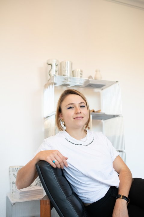 Designerin Yael Anders sitzt auf einem schwarzen Lederstuhl, ein Glasregal im Hintergrund