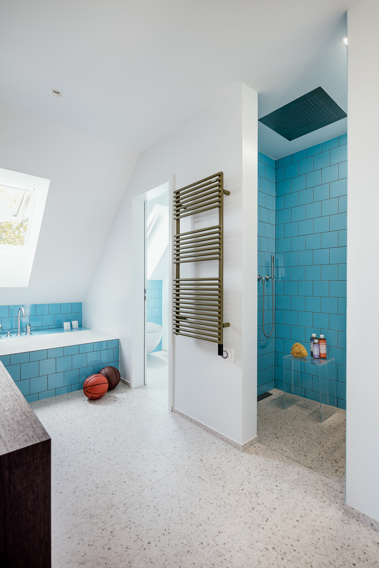 Grosses Badezimmer im Dachgeschoss, heller, gesprenkelter Boden, Duschkabine und Badewanne mit blauen Fliesen, in der Mitte ein Handtuchwärmer an der Wand
