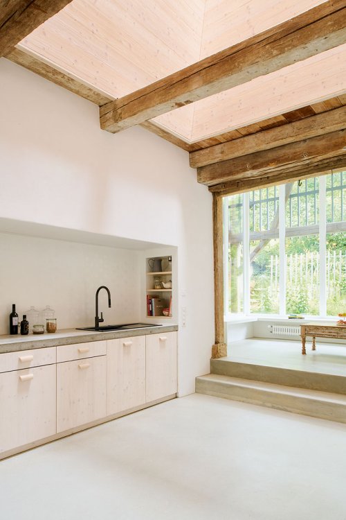 Ein hoher Raum, in dem links eine Küchenzeile steht, daneben blickt man durch ein raumhohes Fenster.