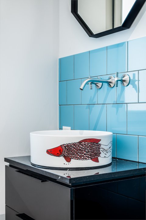 Rundes Waschbecken mit einem roten Fisch darauf auf einem dunklen Möbel, dahinter blaue Fliesen