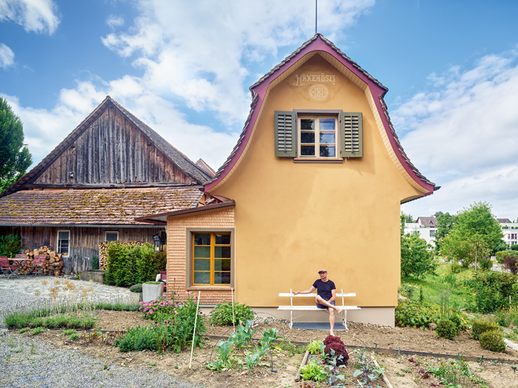 Ein kleines Haus mit gelber Fassade, links davon ein alter Holzbau, vor dem Haus sitzt ein Mann in kurzen Hosen