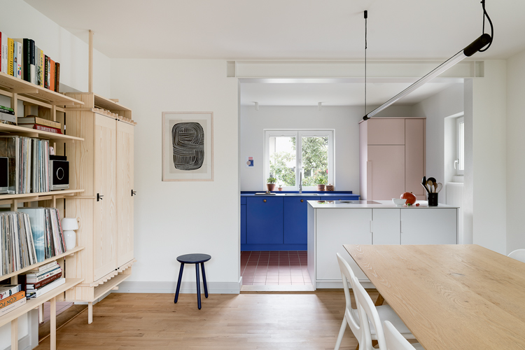 Blick durch ein Esszimmer in eine Küche mit farbigen Fronten, links an der Wand ein schlichtes Holzregal