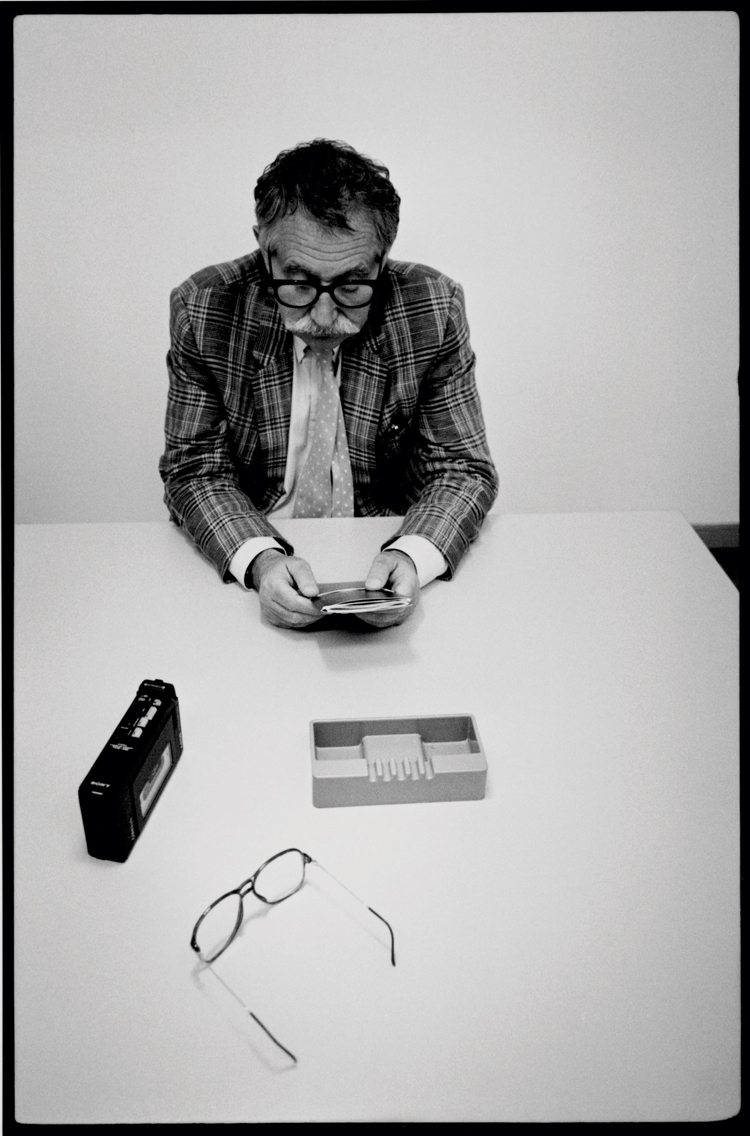 Schwarz-weiss Bild von Designer Ettore Sottsass, von oben fotografiert wie er an einem Schreibtisch sitzt und ein Objekt studiert