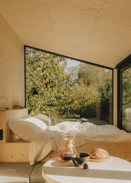 Blick vom innern eines Cabine-House mit hellen Holzwänden, Bett und asymmetrischem Fenster nach draussen in die Natur