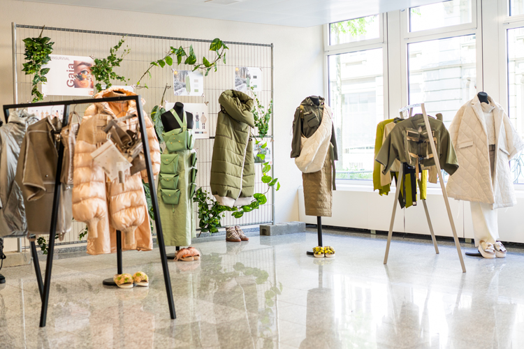 Eingangsbereich der STF wo verschiedene Kleiderständer mit Arbeiten von Studenten ausgestellt sind in den Farben olive, lachs und beige.
