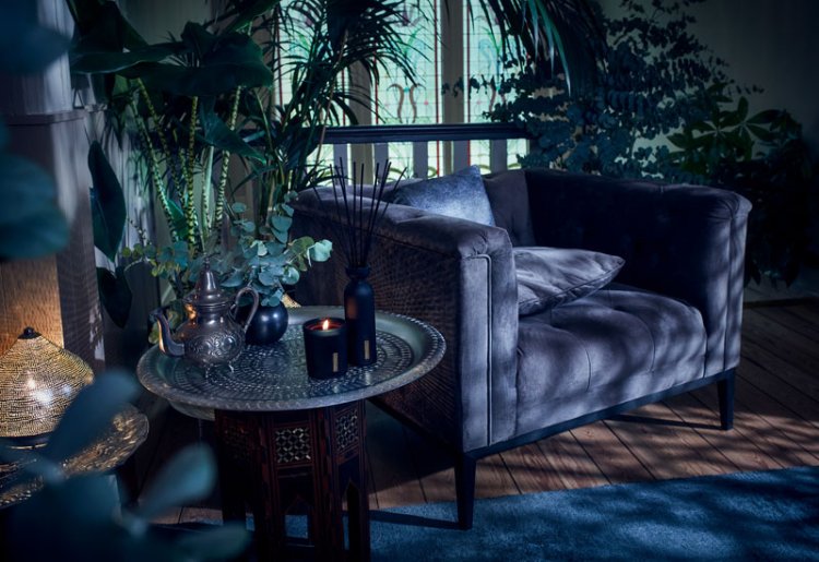Mood-Bild von Rituals, dämmriges Licht, der Blick fällt auf einen dunkel blauen Polstersessel mit einem Sidetable auf der linken Seite wo eine Duftkerze ebenfalls in blau von Rituals steht.