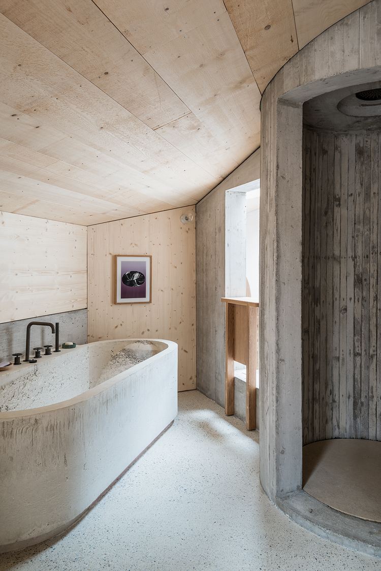 Blick ins Badezimmer mit schrägem Dach aus hellem Holz, links sieht man eine Wanne und rechts eine runde Dusche – beide sind aus Beton gegossen.