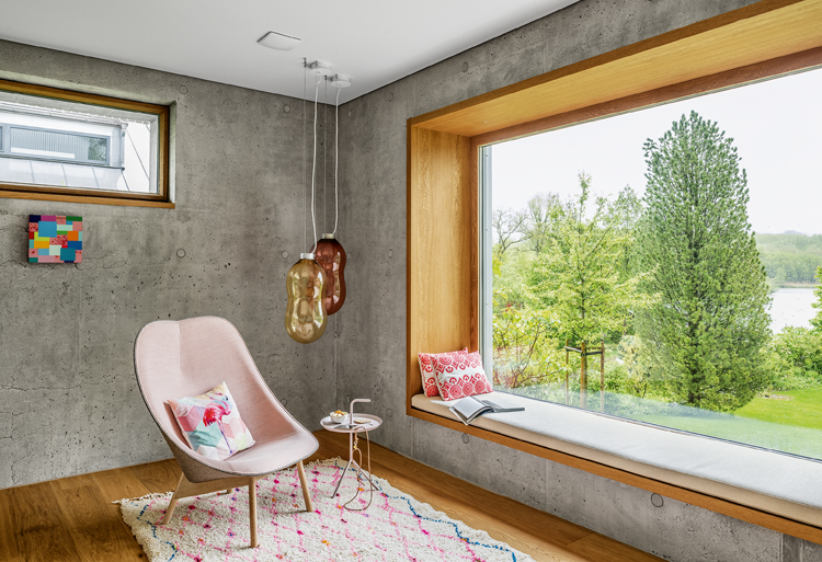 Blick in Zimmerecke, wo sich ein rosafarbiger Sessel befindet, das Fenster zieht sich über die gesamte Front und bietet einen Ausblick ins Grüne. Die Wände sind aus Beton, der Fensterrahmen aus Holz.