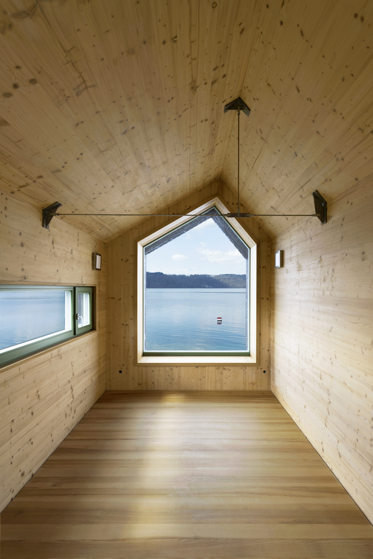 Innenaufnahme eines Tiny Houses am Bodensee, Blick führt durch ein wandgrosses Fenster auf den See, die Innenverkleidung ist minimal und aus hellem Holz.