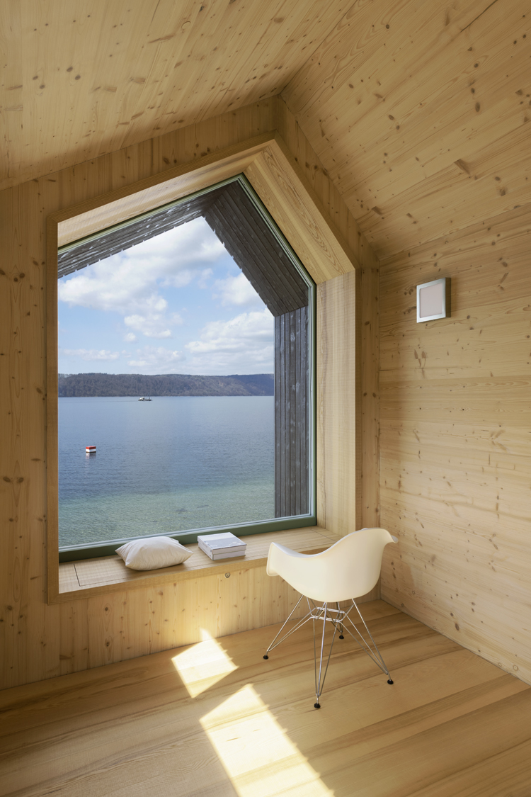 Innenaufnahme eines Tiny Houses am Bodensee, Blick führt durch ein wandgrosses Fenster auf den See, die Innenverkleidung ist minimal und aus hellem Holz.