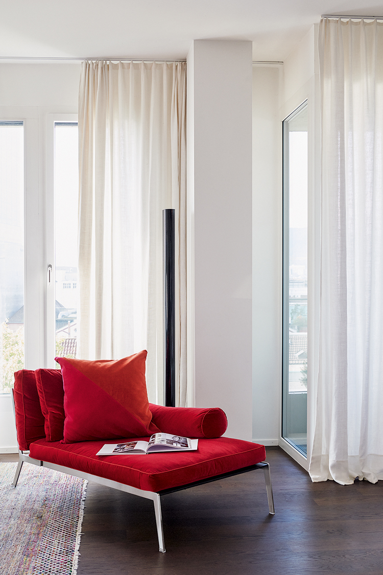 Rote Chaise-Lounge im Wohnzimmer vor weissen Vorhängen.