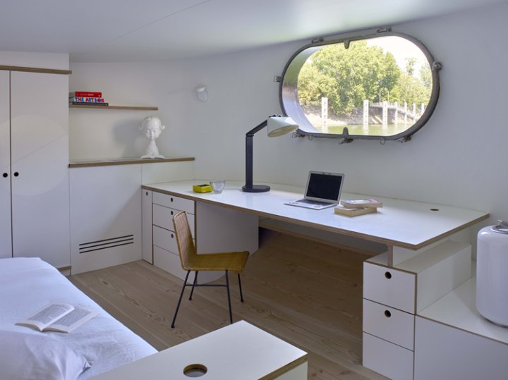 Schlafzimmer auf dem Hausboot in weiss mit Schreibtisch unter dem oval-förmigen Fenster mit Blick auf die Seine.