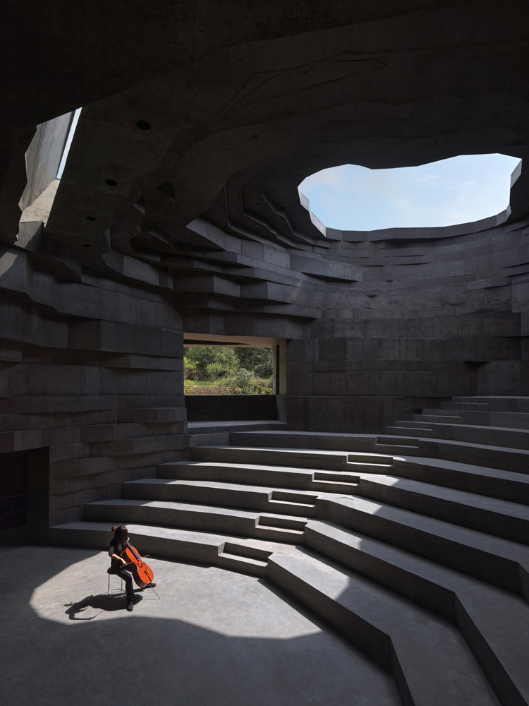 Einsamer Cello-Spieler in einem sonnendurchflutenden Amphitheater mit Betontreppen und organisch geformter Dachöffnung.