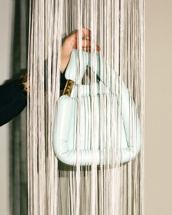 Hellblaue Handtasche wird hinter einem Vorhang aus weissen Fransen in die Luft gehalten.